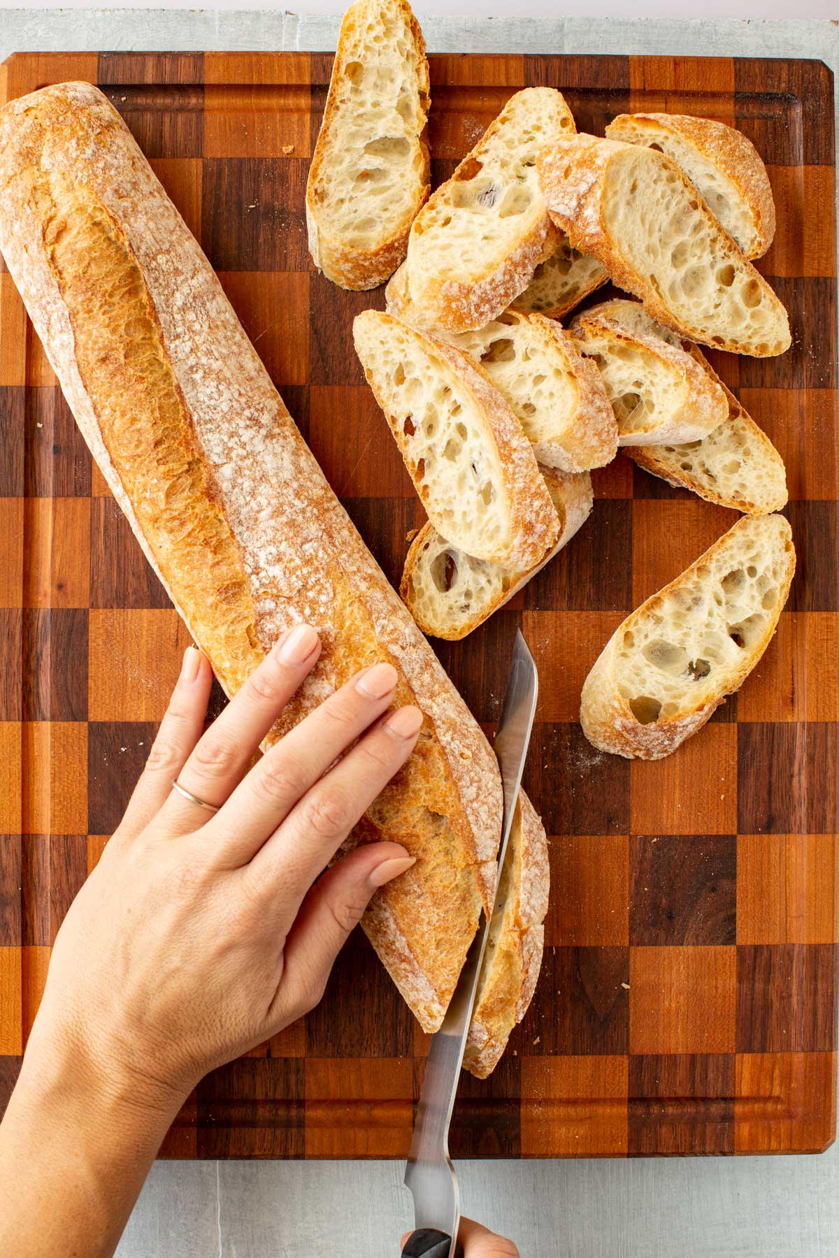 Slicing a fresh baguette on a wooden cutting board with a serrated knife, showing hand placement and even, diagonal slices.
