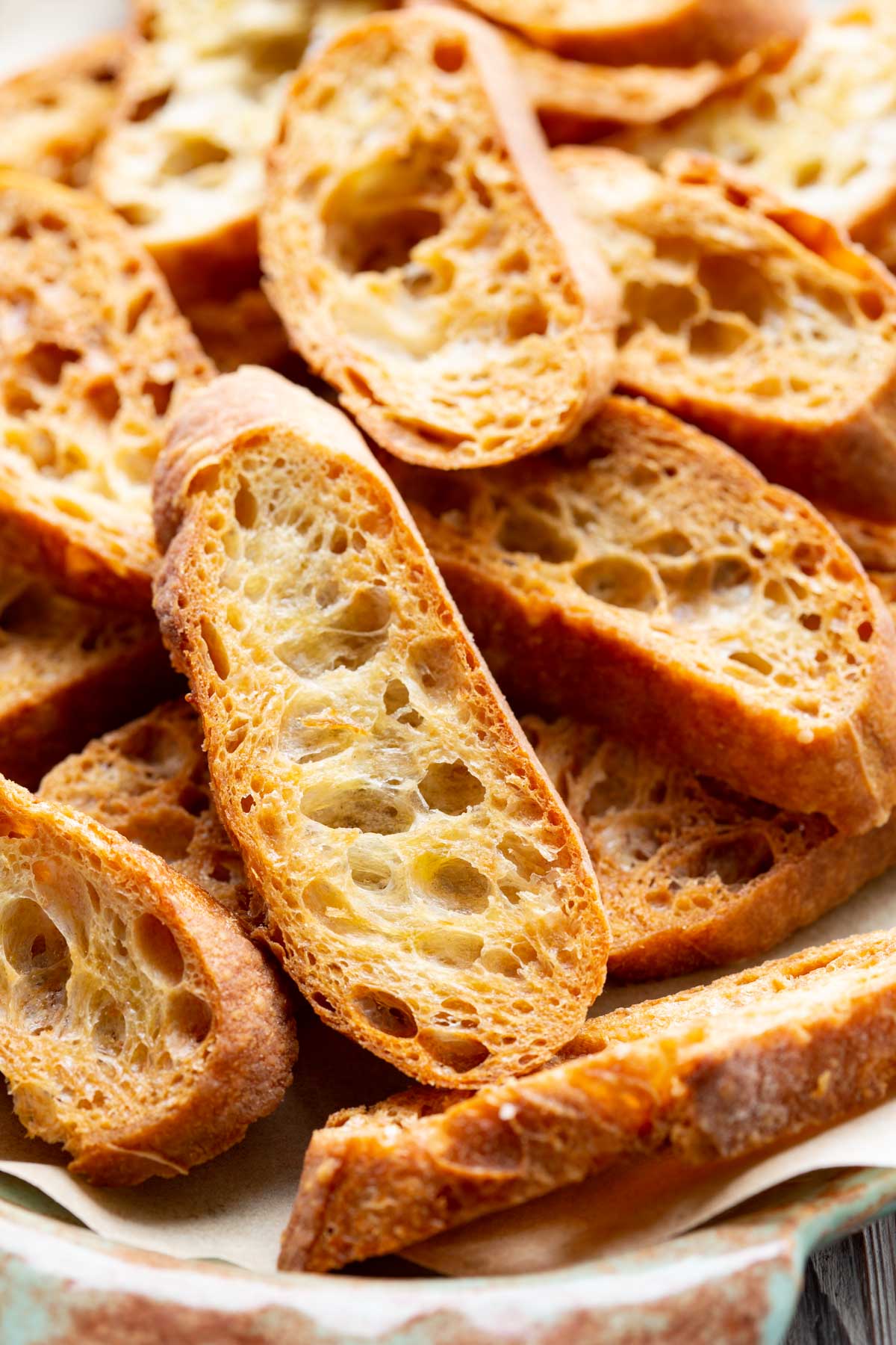 Side angle view of baked crostini stacked on a patterned serving platter lined with parchment paper.