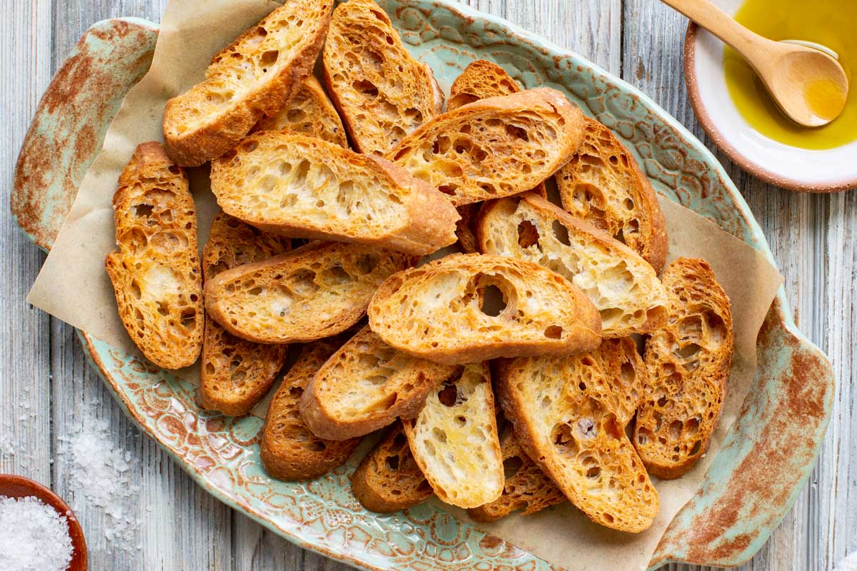 Overhead image of a rustic platter filled with golden crostini beside a small dish of olive oil and sea salt for serving.