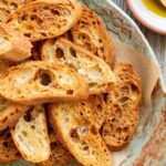 Overhead close-up of golden brown homemade crostini stacked on a rustic green ceramic platter lined with parchment paper, next to a small dish of olive oil with a wooden spoon.