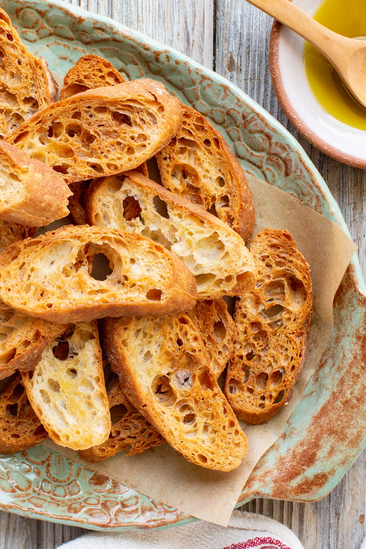 Overhead close-up of golden brown homemade crostini stacked on a rustic green ceramic platter lined with parchment paper, next to a small dish of olive oil with a wooden spoon.