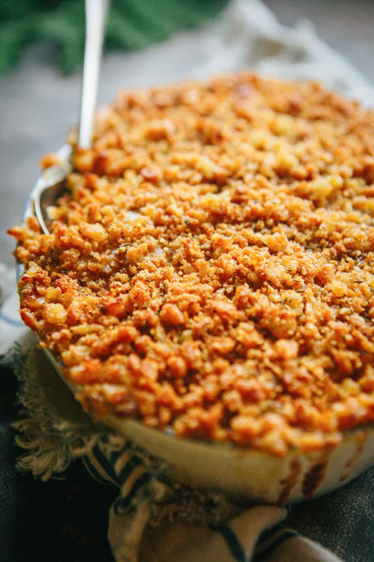 Close-up of creamy kale au gratin with a crisp, golden breadcrumb topping in a round baking dish, with a serving spoon tucked into the side.