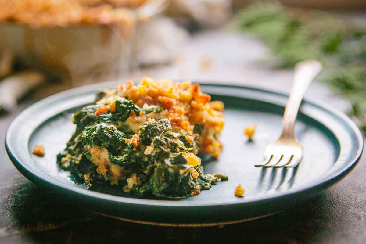 A single serving of creamy kale au gratin on a dark plate, showing tender greens coated in sauce and topped with toasted breadcrumbs.