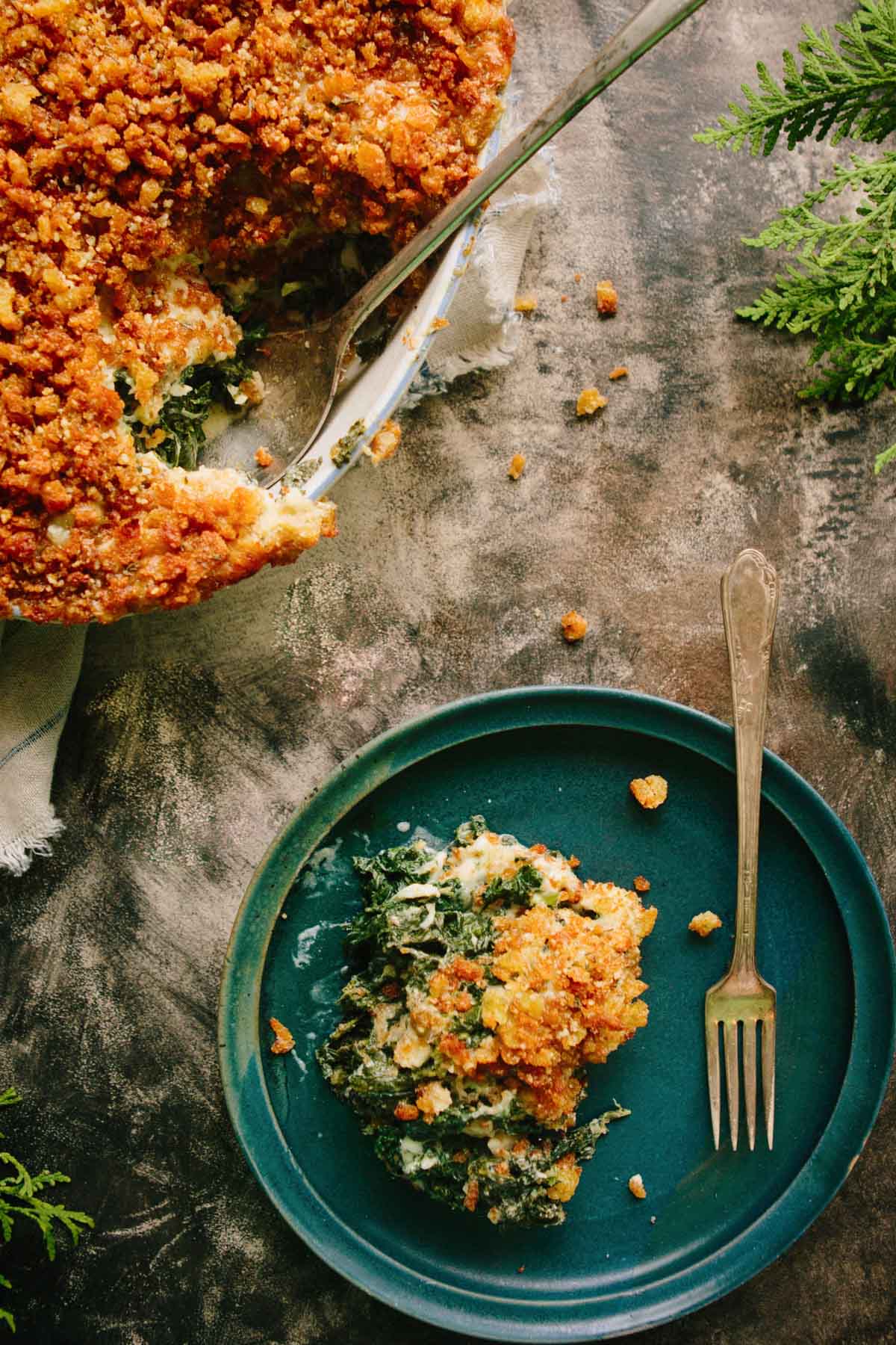 Overhead shot of a kale gratin with a slice removed, revealing creamy greens beneath the crunchy golden topping, served next to a plated portion with a fork.