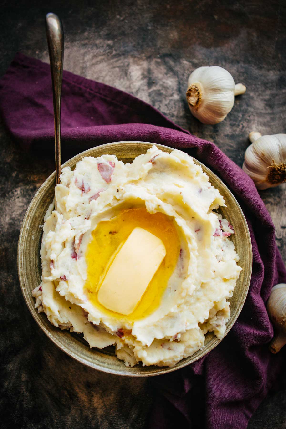 Overhead view of mashed potatoes in a rustic bowl topped with a pat of butter, surrounded by garlic bulbs and a dark napkin.