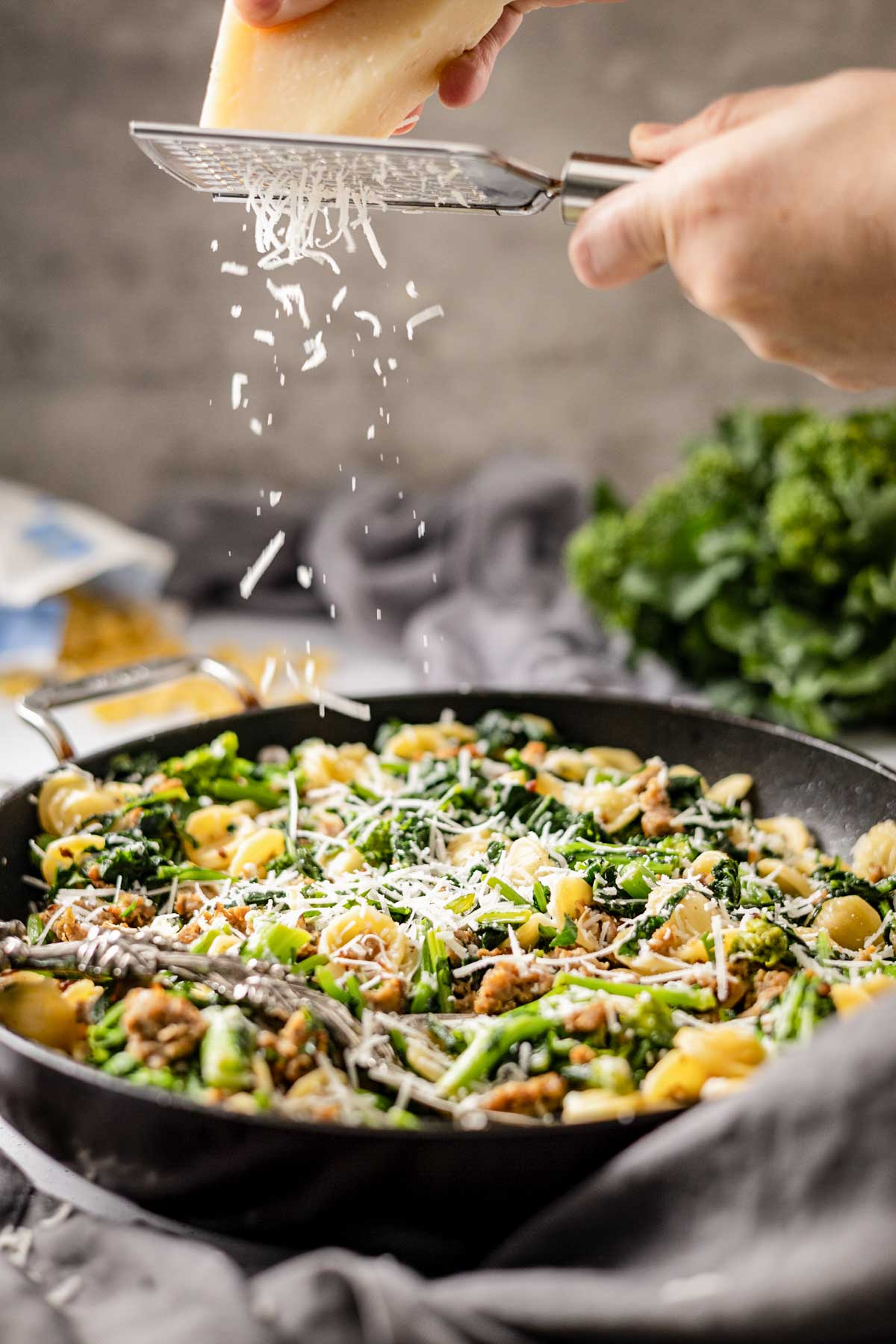 Hands grating Parmigiano Reggiano cheese over a skillet of orecchiette with sausage and greens.