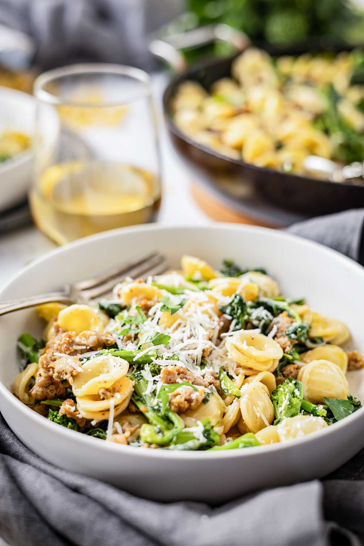 Bowl of orecchiette with broccoli rabe and sausage set on a table with a glass of white wine in the background.