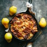 Pear pandowdy in a black cast iron skillet surrounded by fresh yellow pears on a dark surface.