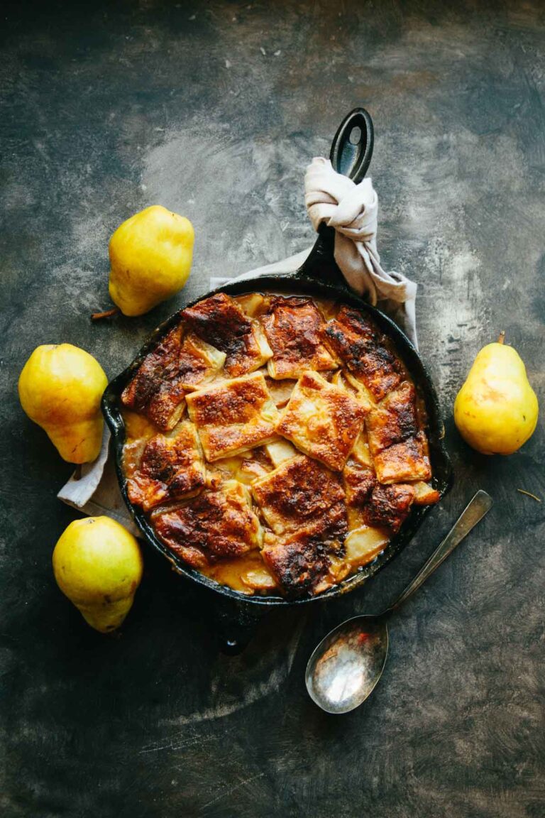 Pear pandowdy in a black cast iron skillet surrounded by fresh yellow pears on a dark surface.