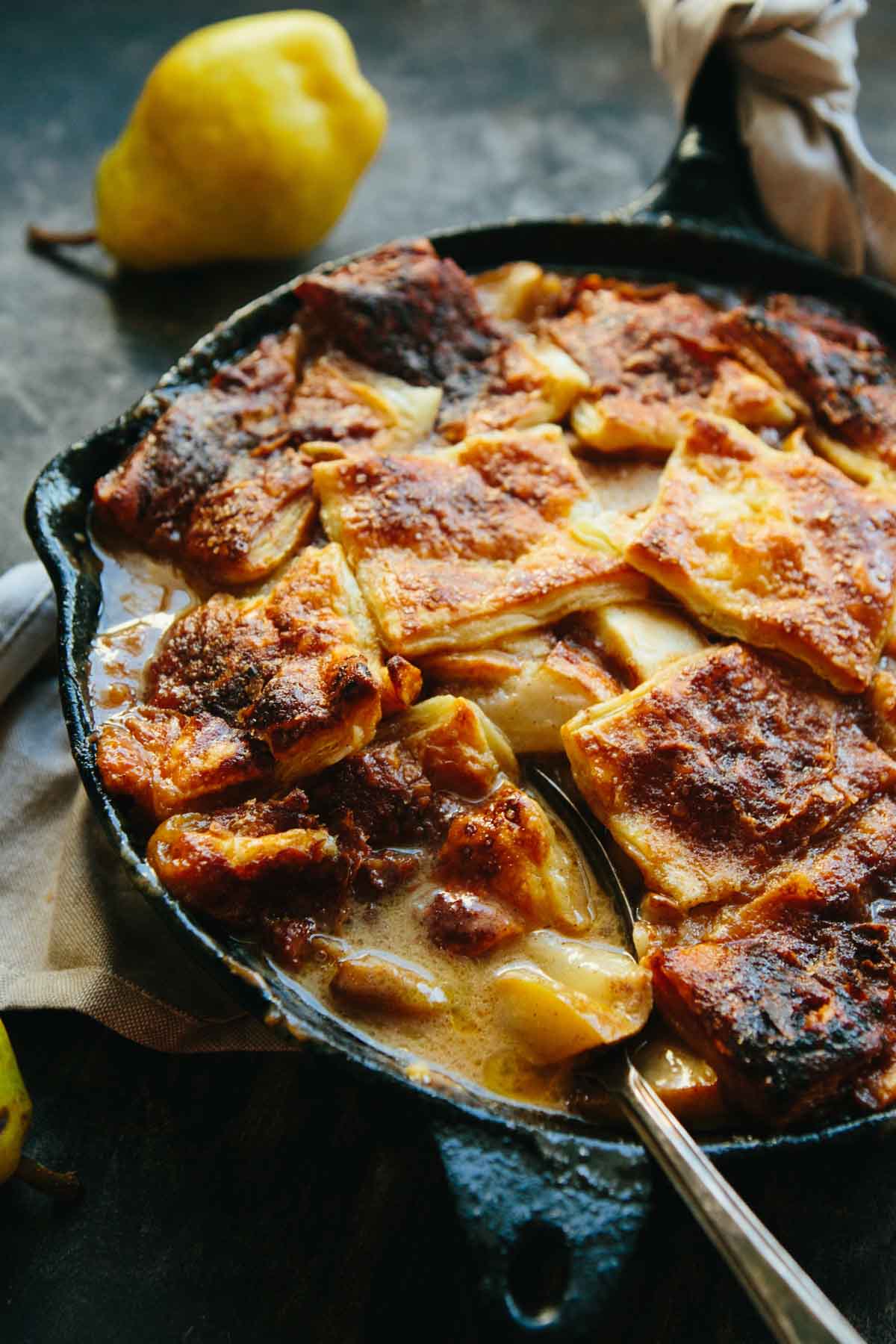 Warm pear pandowdy in a skillet with visible layers of flaky puff pastry and a spoon tucked inside.