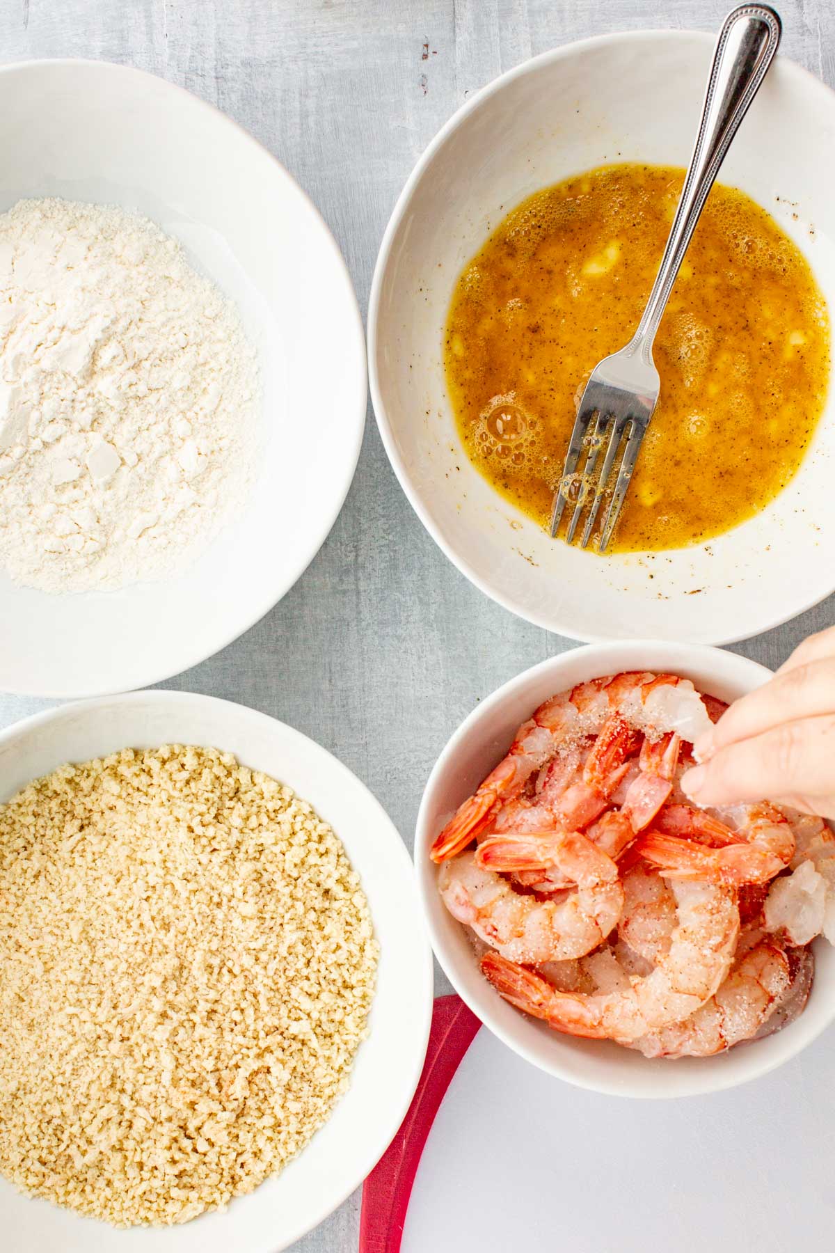 Overhead shot of separated ingredients for fried shrimp including raw shrimp, flour, panko breadcrumbs, cayenne, lemon wedges, egg, oil, salt, and pepper in small glass bowls on a gray surface.