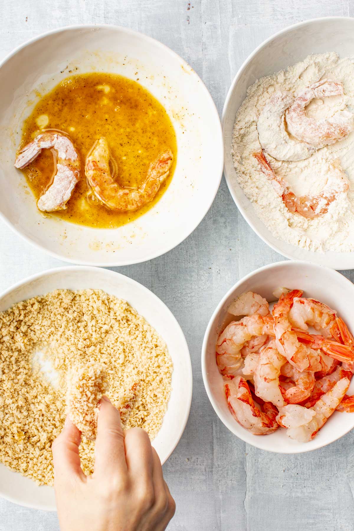 Setup of breading station with bowls of flour, beaten egg, and panko breadcrumbs next to a bowl of raw shrimp being seasoned.