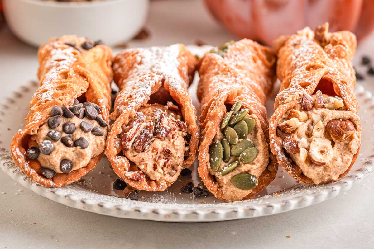 Four pumpkin cannoli on a white plate, each filled with creamy pumpkin ricotta filling and topped with chocolate chips, nuts, and pumpkin seeds.
