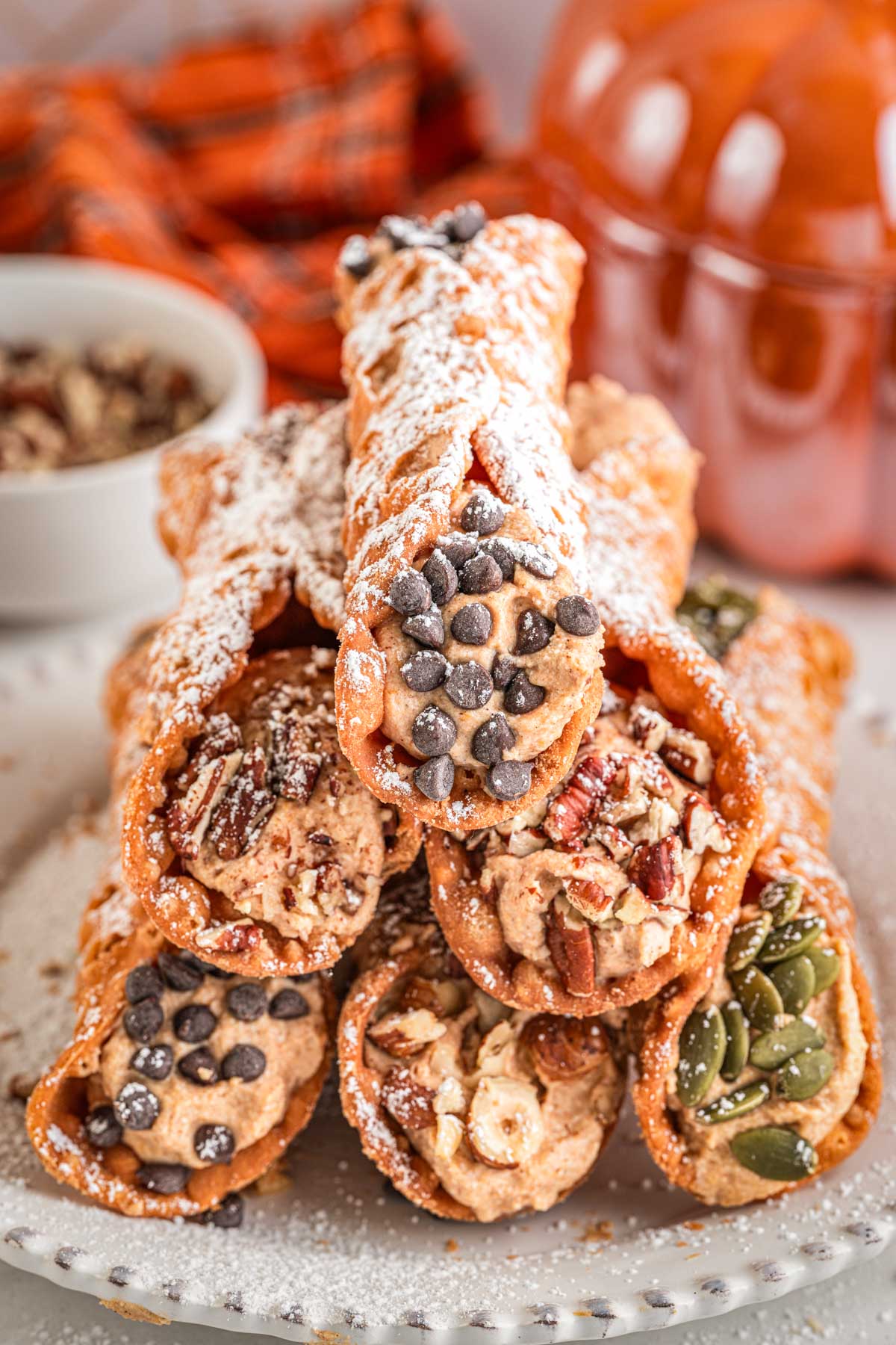 Stack of assorted pumpkin cannoli topped with mini chocolate chips, crushed pecans, and pumpkin seeds, dusted with powdered sugar.