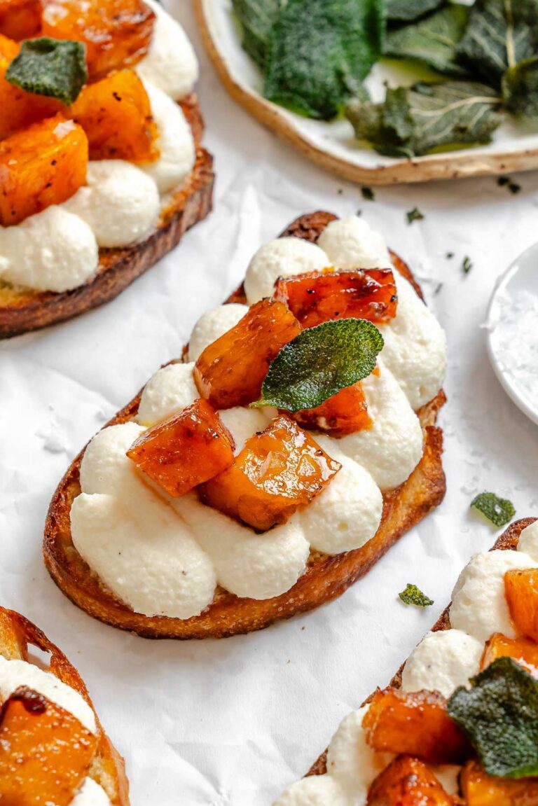 Finished pumpkin ricotta crostini topped with caramelized squash and fried sage leaves arranged on parchment paper.
