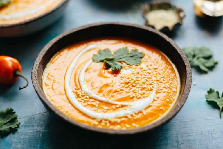 Bowl of spicy carrot soup garnished with sesame seeds and cilantro, photographed at an angle with habanero peppers in the background.
