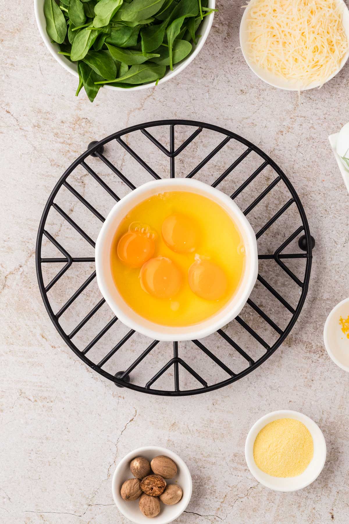 Four raw eggs in a white bowl on a black trivet, surrounded by small bowls of nutmeg, semolina, lemon zest, grated Parmesan, and a larger bowl of fresh spinach.