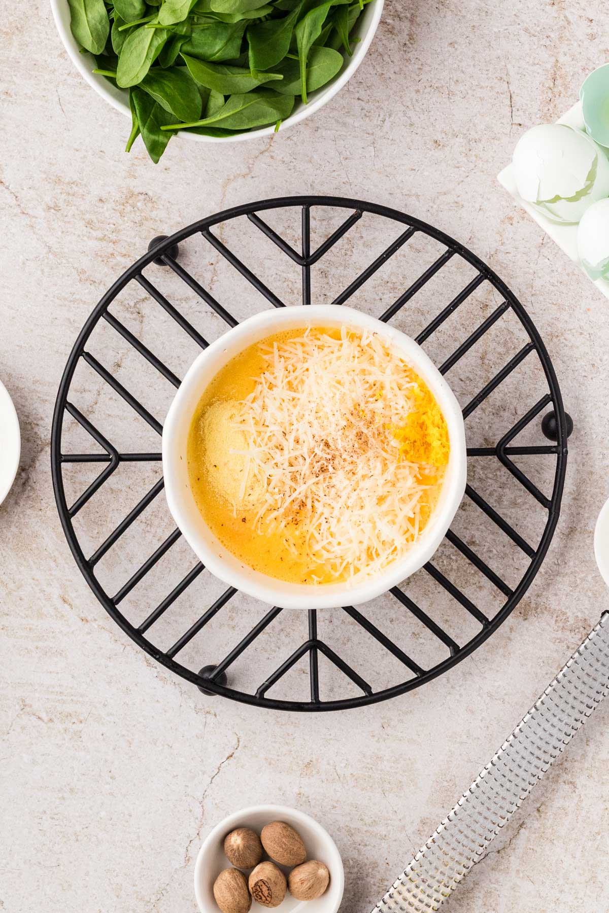 Eggs whisked with Parmesan, nutmeg, lemon zest, and semolina in a white bowl, with fresh spinach and egg shells nearby.