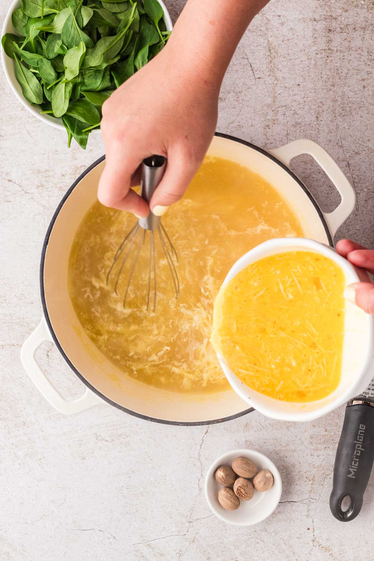 Hand pouring the egg and cheese mixture into a simmering pot of broth while whisking to form delicate shreds of egg.