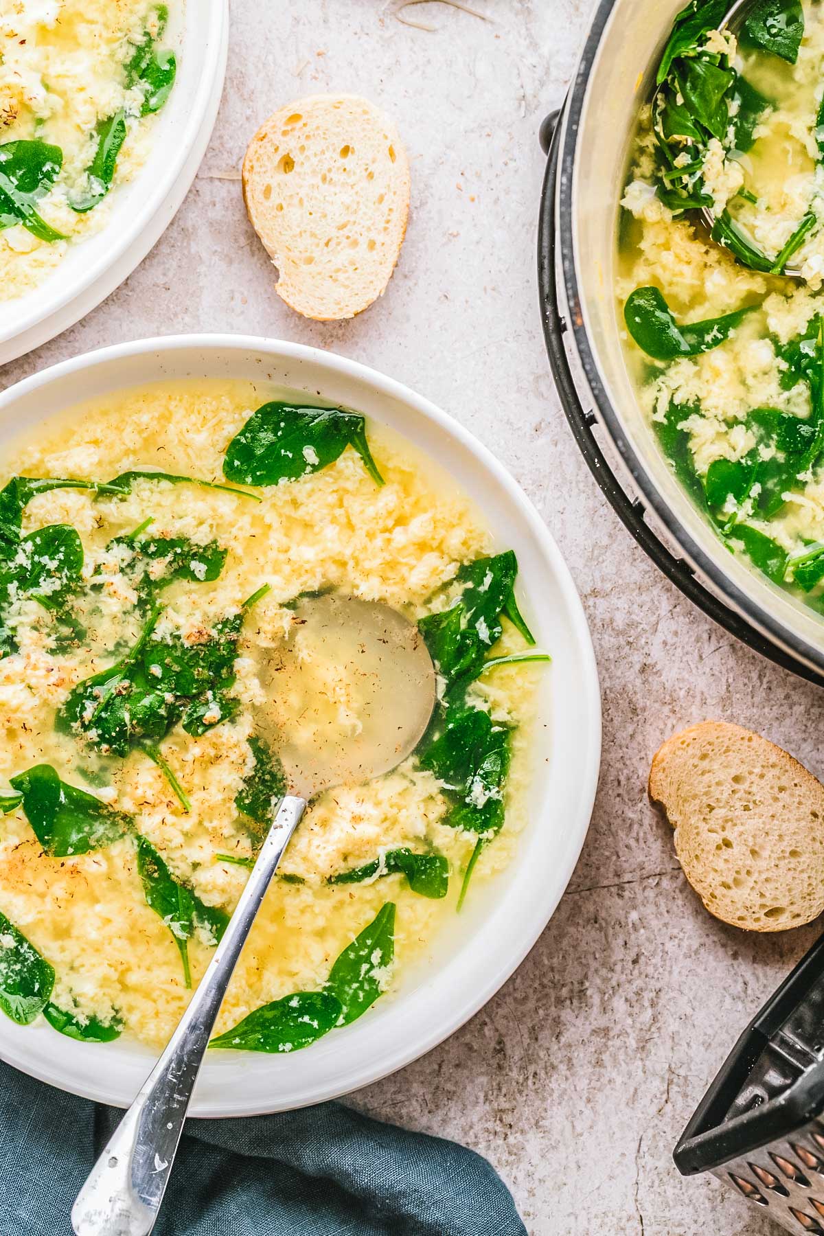 Close-up of stracciatella soup ladled into white bowls, with spinach and egg strands visible, served with slices of bread.