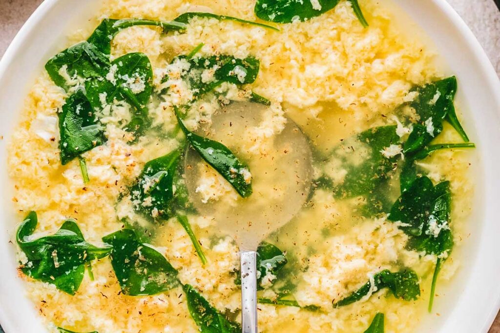 Zoomed-in view of stracciatella soup showing fluffy shreds of egg and tender spinach leaves in a clear broth.