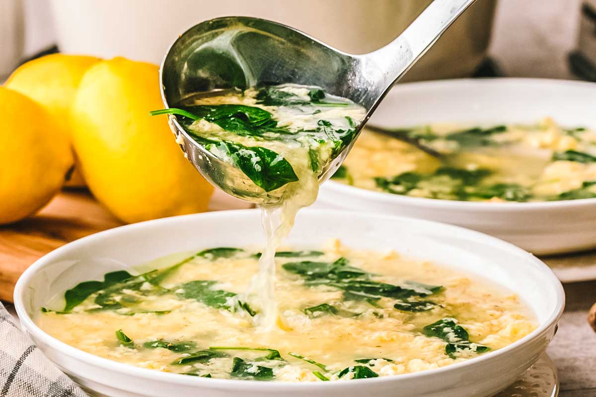 A ladle lifting stracciatella soup from a white bowl, showing silky egg strands and spinach against a backdrop of lemons.