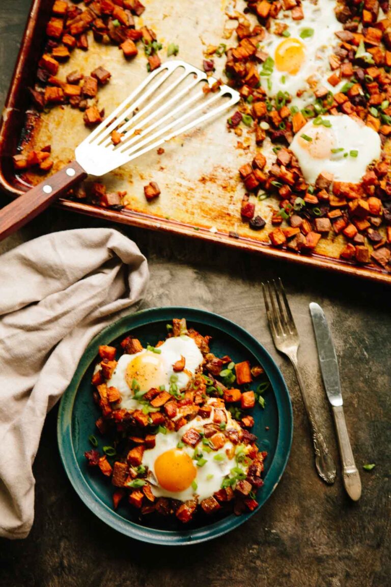 Overhead view of a baking sheet filled with roasted sweet potatoes, chorizo, and baked eggs, with a serving plate nearby.