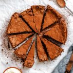 Overhead shot of sliced Torta Caprese dusted generously with cocoa powder, arranged on parchment with a bowl of whipped cream and a sieve nearby.