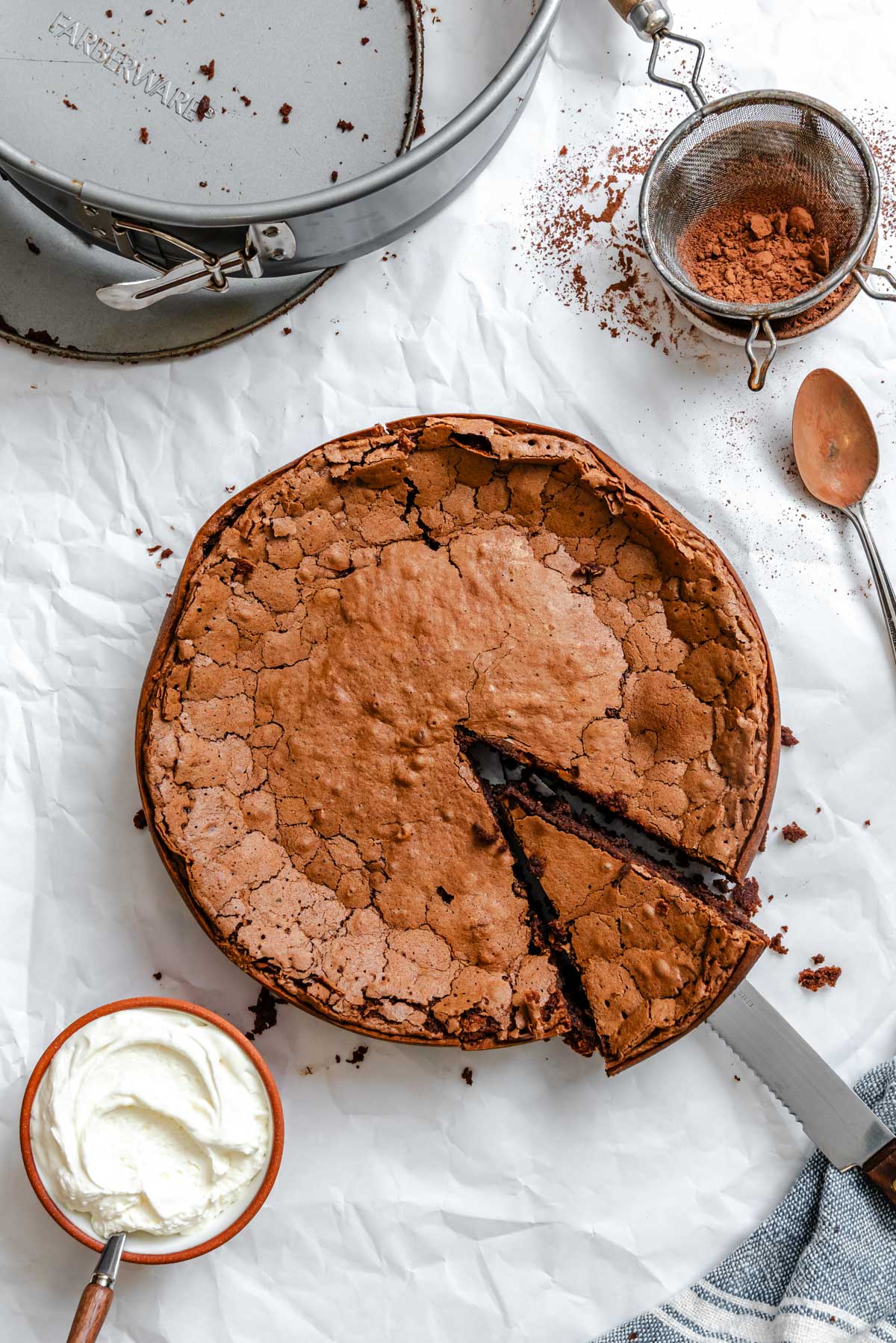 Whole Torta Caprese on parchment with one slice removed, showing the glossy, crackled crust beside a bowl of whipped cream and a metal sieve with cocoa powder.