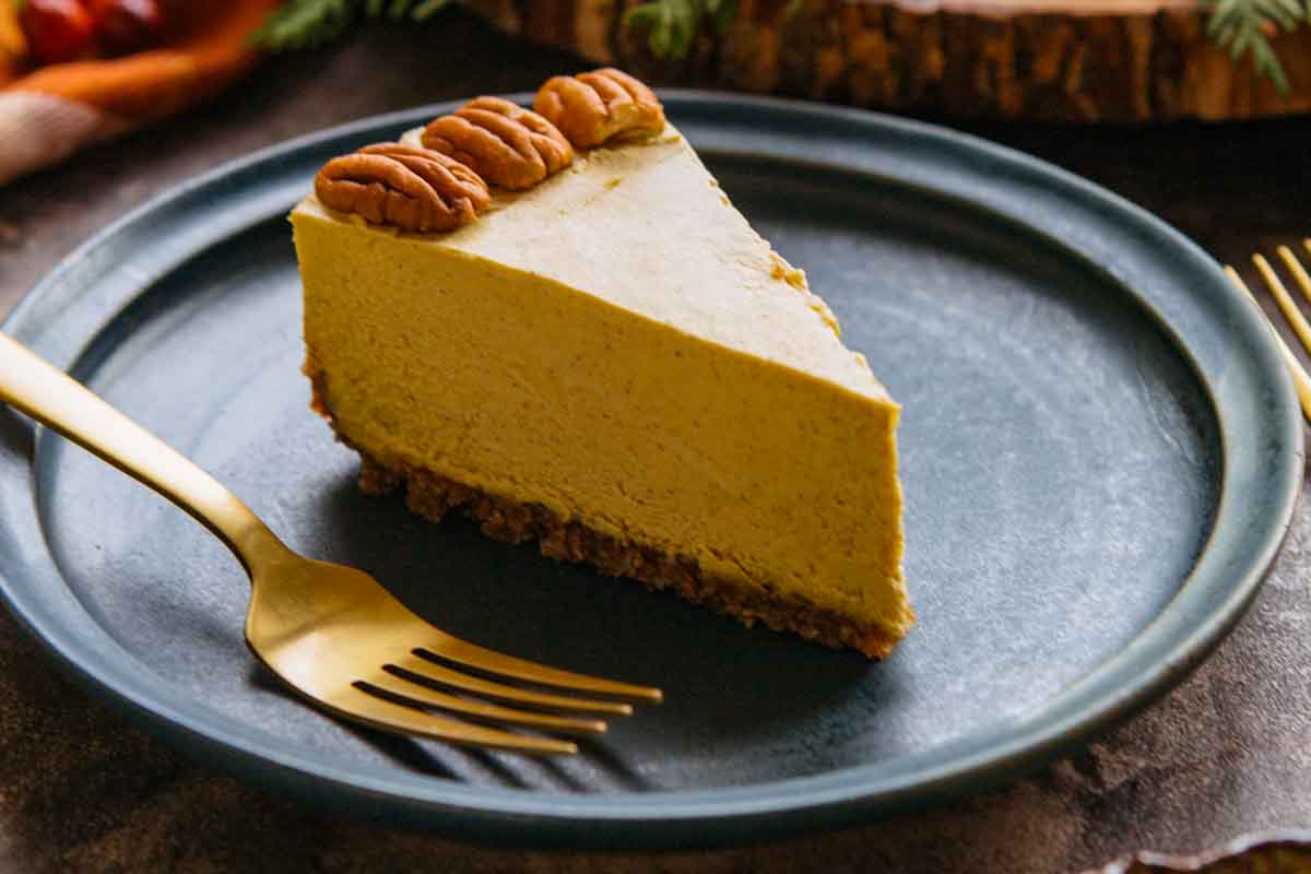 Slice of vegan pumpkin cheesecake on a dark blue plate with a gold fork, showing its creamy texture and nut crust.