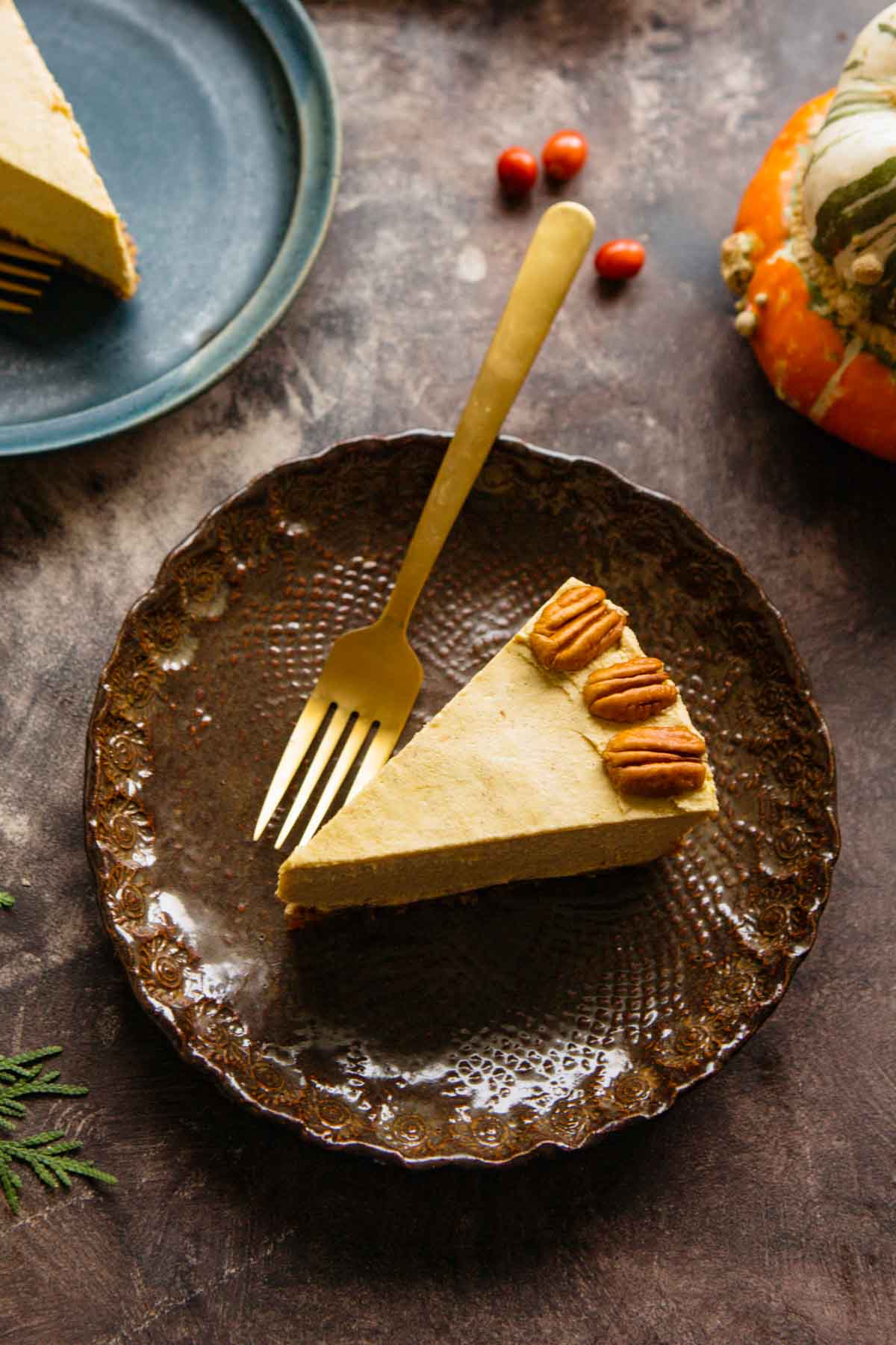 Slice of vegan pumpkin cheesecake on a brown plate with a gold fork beside it and a small pumpkin in the background.