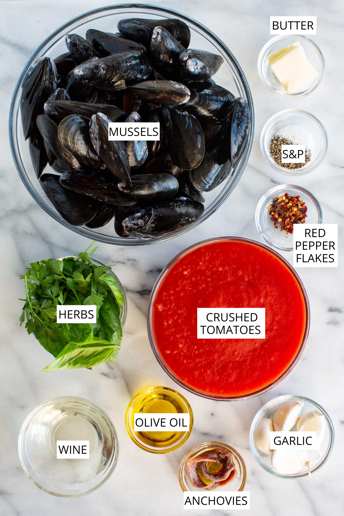 A top down image showing labeled mussels, crushed tomatoes, garlic, anchovies, olive oil, wine, butter, herbs and seasonings arranged on a marble surface.