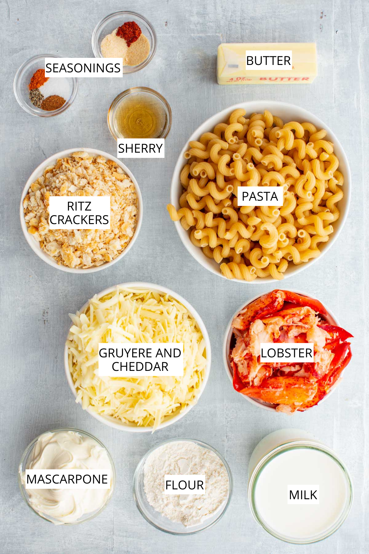 A flat lay of all ingredients for lobster mac and cheese including lobster, pasta, Ritz crackers, shredded cheese, mascarpone, milk, butter, flour, sherry, and seasonings, each labeled.