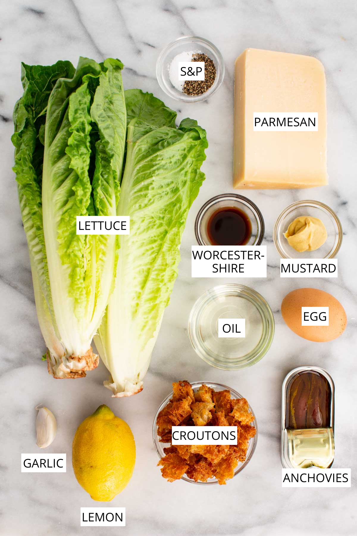 Overhead layout of the main Caesar salad ingredients on marble, including romaine lettuce, parmesan, egg, anchovies, garlic, lemon, oil, Worcestershire sauce, mustard, salt and pepper and croutons.