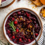 A serving bowl of glossy balsamic cranberry sauce topped with fresh orange zest and a sprig of rosemary, surrounded by cranberries and zest on the table.