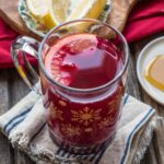 Blueberry Ginger Hot Toddy in a snowflake glass mug garnished with a lemon slice, sitting on a striped cloth napkin beside lemon wedges and honey.