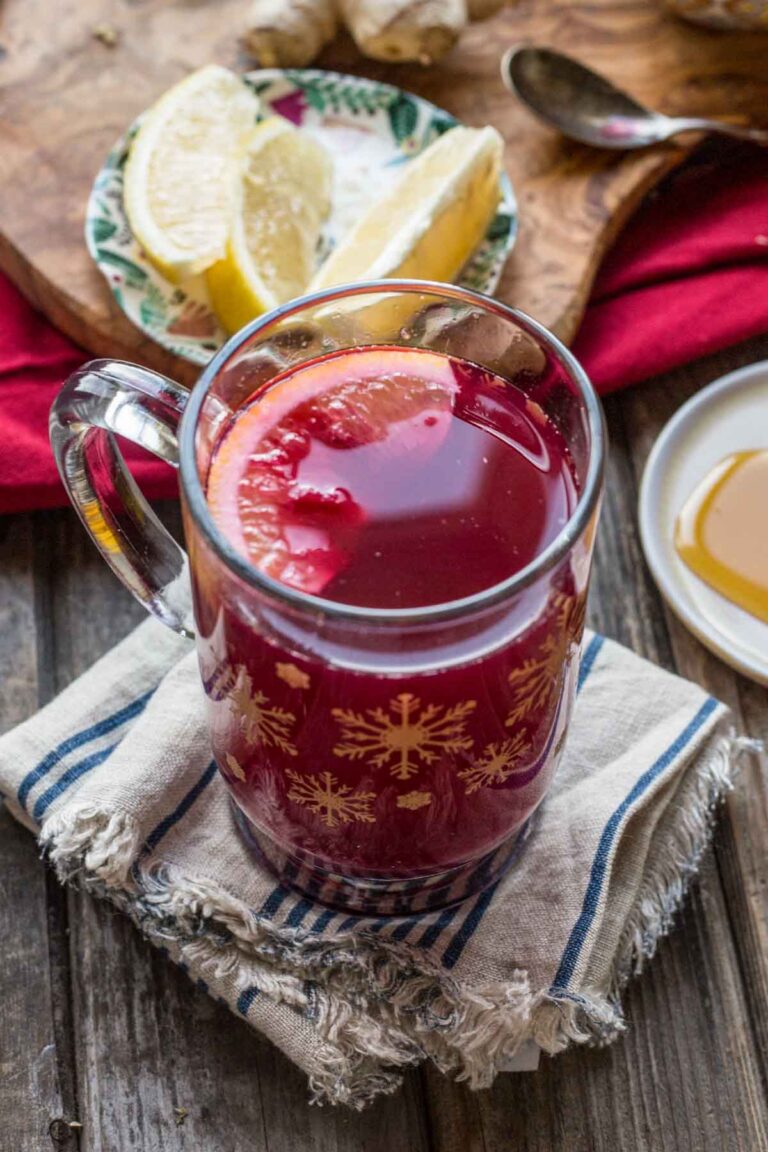 Blueberry Ginger Hot Toddy in a snowflake glass mug garnished with a lemon slice, sitting on a striped cloth napkin beside lemon wedges and honey.