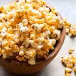 Wooden bowl filled with honey butter buffalo popcorn with pieces scattered on parchment.