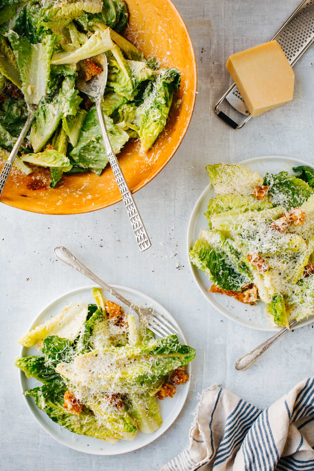 Overhead photo showing a large wooden bowl of Caesar salad with two smaller plated servings and a block of parmesan on a grater nearby.