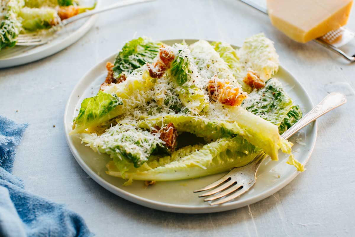 Side angle of a plated Caesar salad with romaine, croutons and grated parmesan, with a fork resting on the plate.