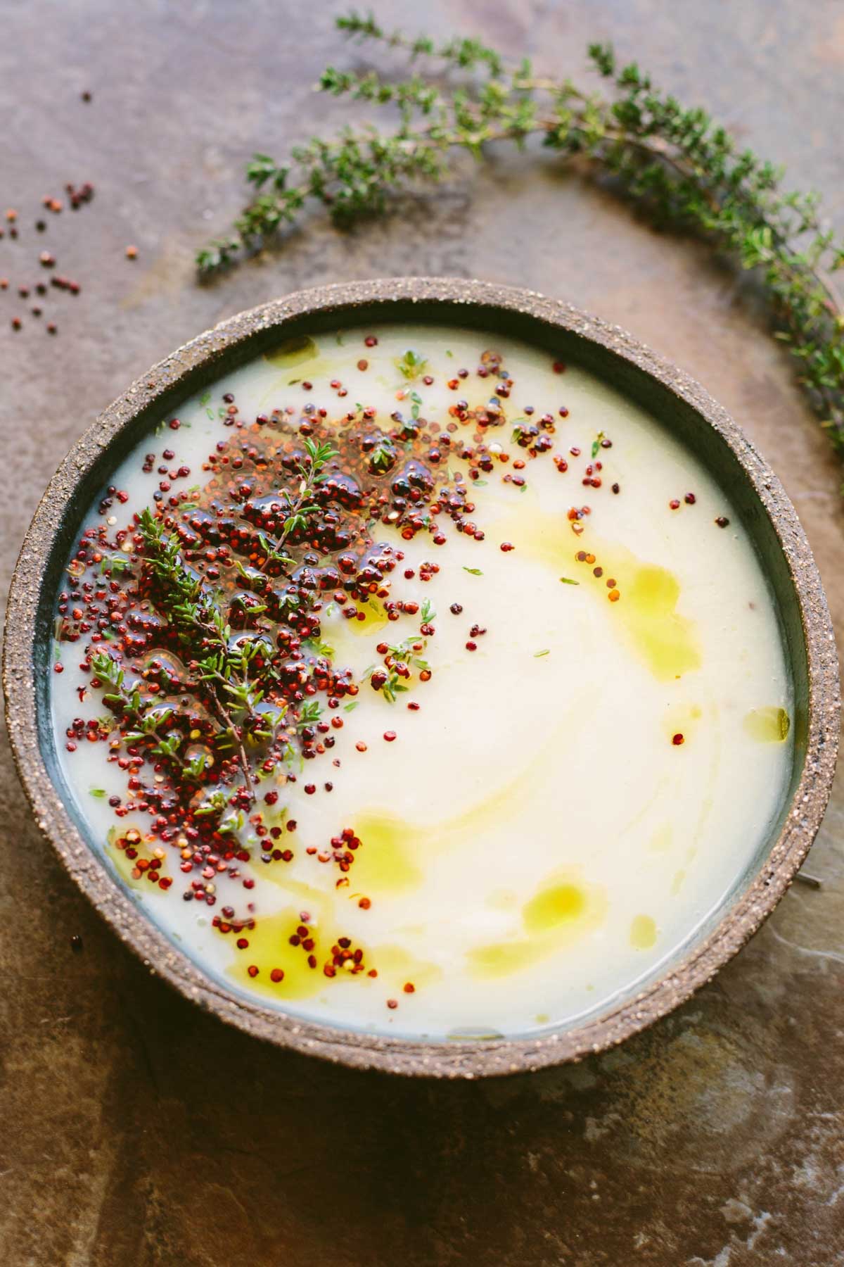 A creamy white bowl of parsnip cauliflower soup topped with puffed red quinoa, fresh thyme sprigs and a drizzle of olive oil.