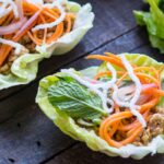 Fresh lettuce wraps filled with seasoned chicken, pickled carrots, herbs and crispy puffed rice noodles on a wooden surface.