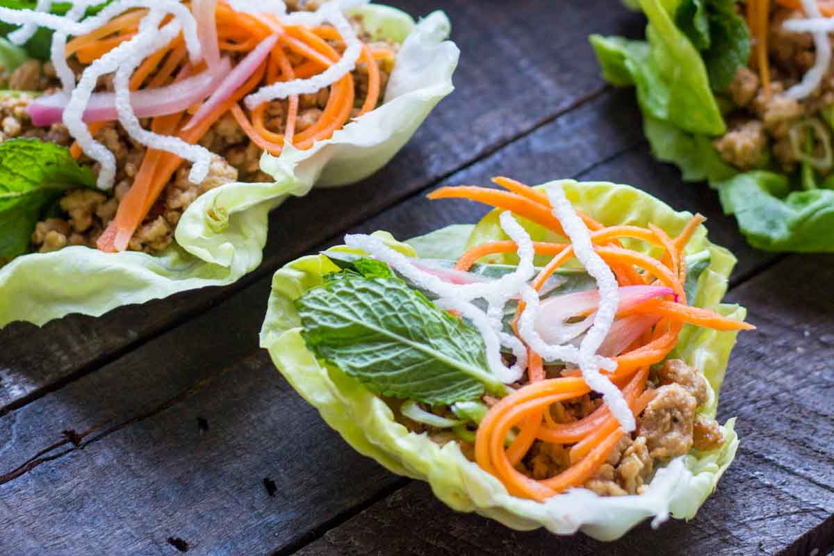 Fresh lettuce wraps filled with seasoned chicken, pickled carrots, herbs and crispy puffed rice noodles on a wooden surface.