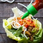 A lettuce wrap filled with chicken, herbs, pickled vegetables and puffed noodles being drizzled with Sriracha sauce.