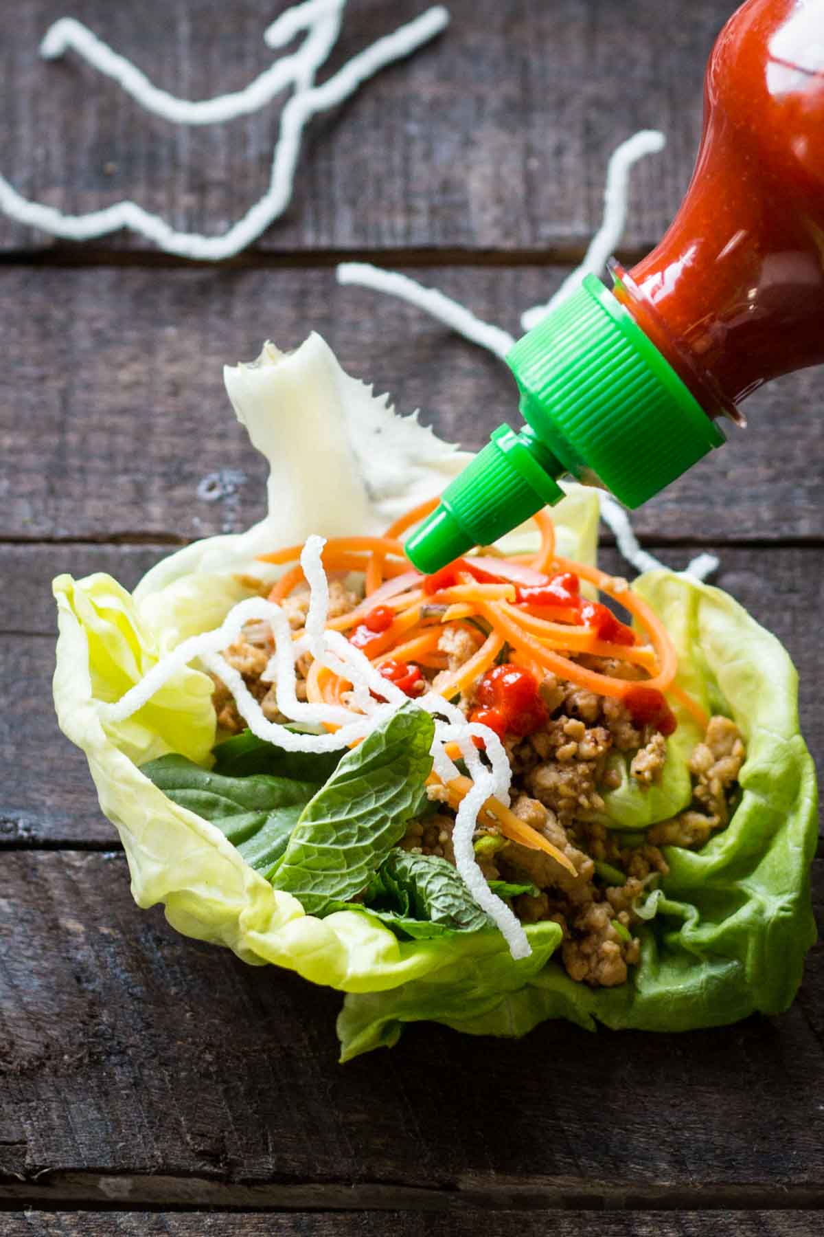 A lettuce wrap filled with chicken, herbs, pickled vegetables and puffed noodles being drizzled with Sriracha sauce.