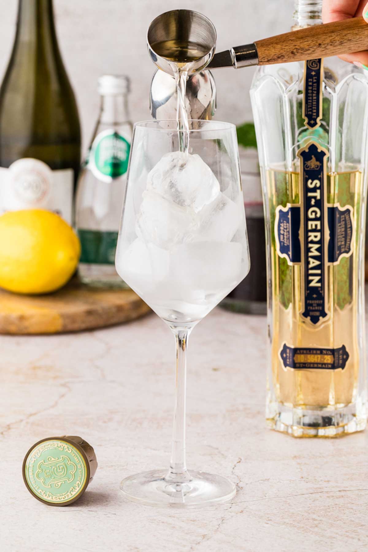 Pouring elderflower liqueur into a wine glass filled with ice cubes, with lemons, prosecco, and mint in the background.