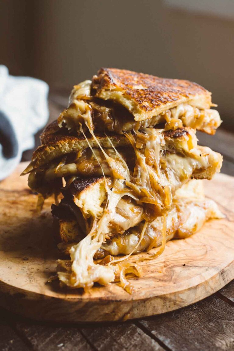 Stack of French Onion Grilled Cheese sandwiches on a wooden board, filled with gooey melted cheese and caramelized onions spilling out.
