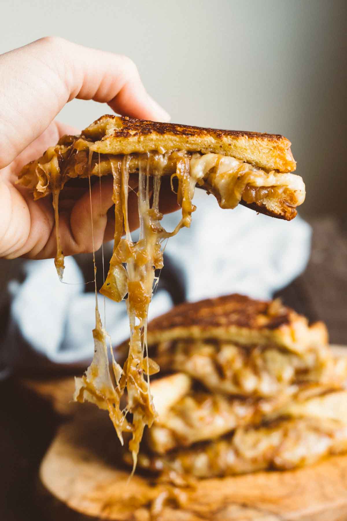 Hand holding a slice of French Onion Grilled Cheese with melted cheese stretching and onions dripping from the edges.