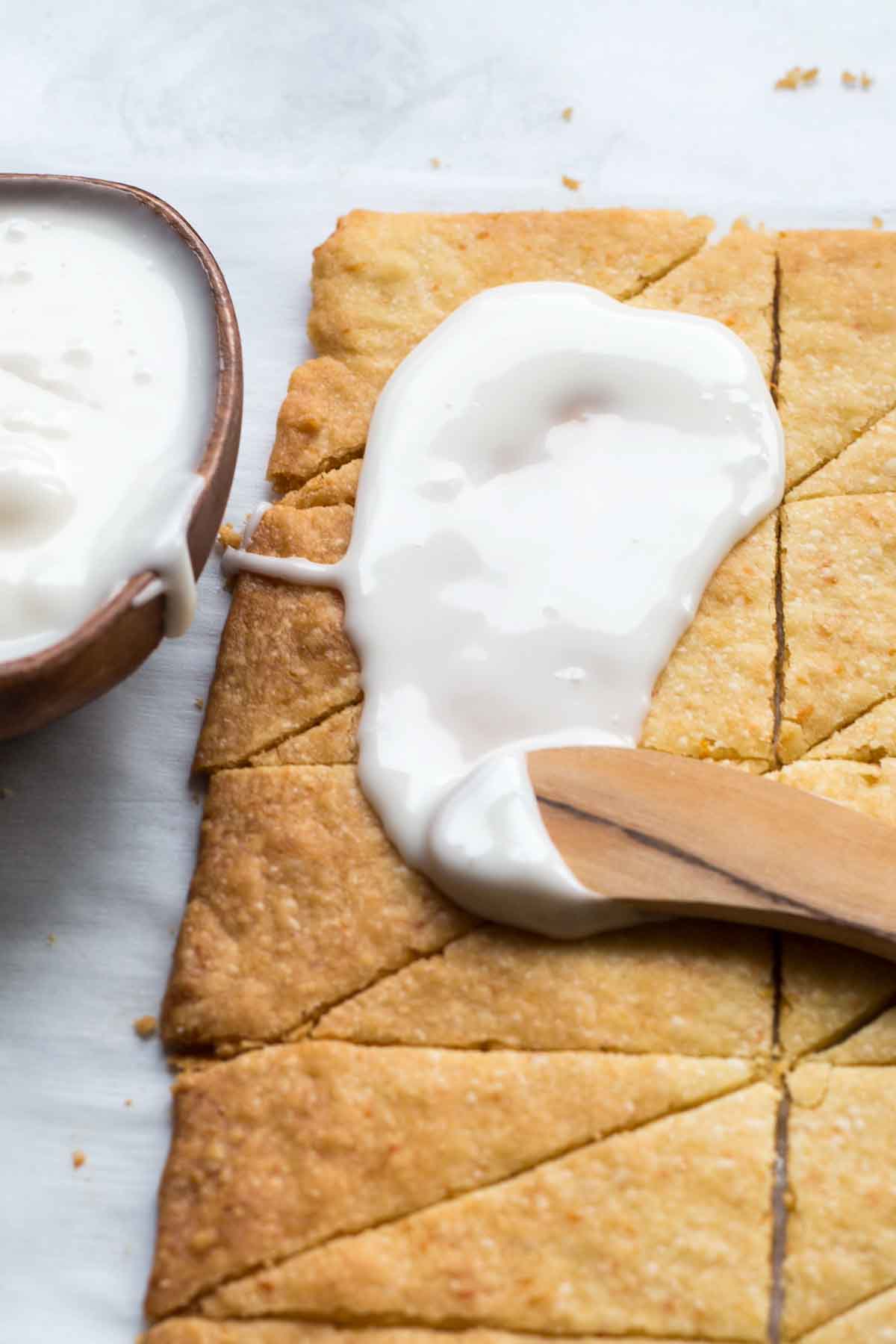 A close up of warm shortbread cut into triangles with a thin layer of white grapefruit glaze being spread over the surface using a small wooden spatula.