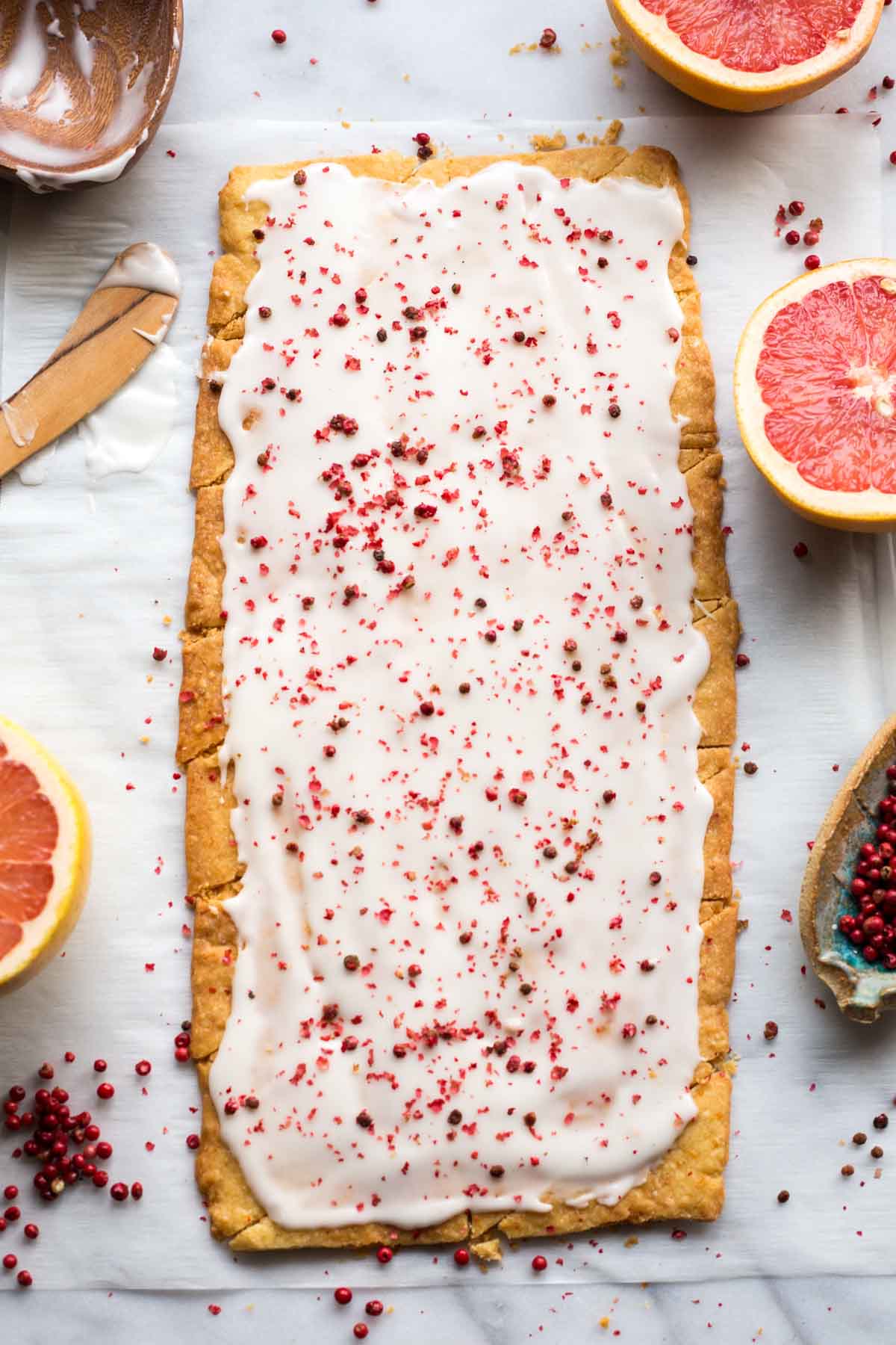 A full sheet of glazed grapefruit shortbread topped with crushed pink peppercorns, surrounded by halved grapefruit and a wooden bowl with extra icing.